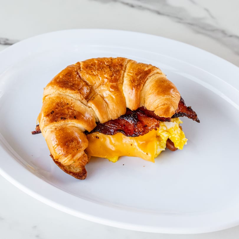 A golden croissant breakfast sandwich with bacon, scrambled eggs, and melted cheese on a white plate.