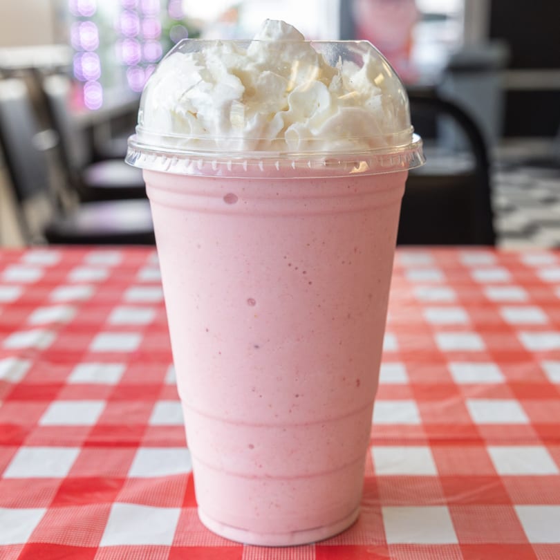 Pink strawberry milkshake with whipped cream in a plastic cup on a red and white checkered tablecloth.