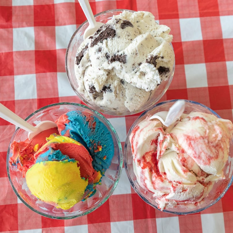 Three bowls of ice cream on a red and white checkered tablecloth, including cookies and cream, rainbow, and strawberry swirl.