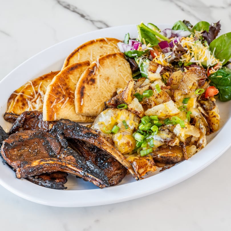 Grilled lamb chops, cheesy potatoes, fresh salad, and flatbread on a white plate.